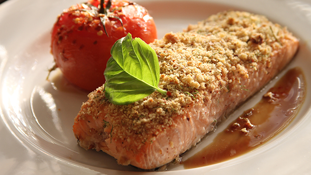 This is an image of a baked breaded piece of salmon on a white plate with a garnish and a baked whole tomato.