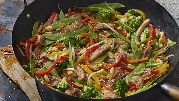 This is an image of a wok containing sliced red and green peppers, snap peas, sliced white onion, broccoli, peanuts, sesame seeds, sliced carrots, and bean sprouts.