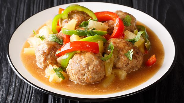 This is an image of a white bowl containing cooked meatballs, sliced red and green peppers, and cubed pineapple in a liquid sauce.