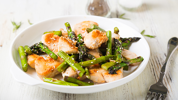 Cubed cooked chicken with pieces of cooked asparagus combined on a white plate with a silver fork next to it