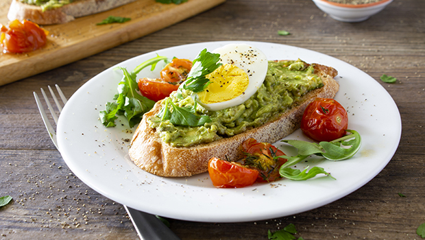 Sliced piece of bread with smashed avocado spread and sliced half of a hard-boiled egg on top with cooked cherry tomatoes all on a white plate