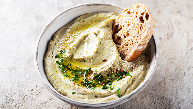 White bowl filled with an eggplant hummus spread with an olive oil drizzle and a sliced piece of bread