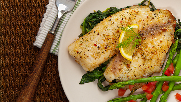Baked fish on a white plate with cooked spinach, asparagus, and diced tomatoes next to a fork