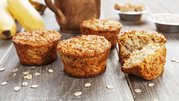 Four banana muffins on a wooden table, with one muffin cut in half