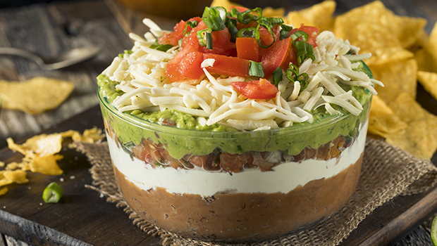 Glass dish with layers of bean dip, sour cream, pico de gallo, guacamole, and cheese with diced tomatoes on top