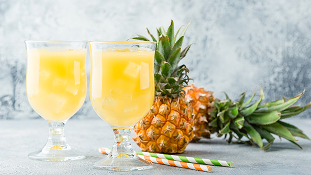 Two glasses of pineapple juice with ice cubes, two straws, and two whole pineapples on a backdrop