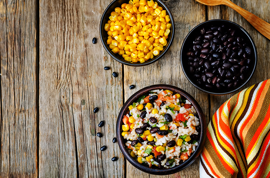 Black beans, rice and con mixed in a black bowl with a black bowl full of corn and another bowl full of black beans, all on a wooden table